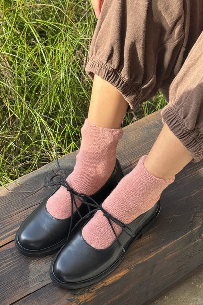 Person wearing black shoes and pink socks sitting on a wooden bench with grass in the background