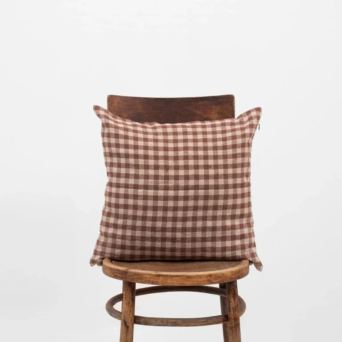 A square throw pillow with a brown and beige gingham check pattern, displayed on a rustic wooden stool against a plain white background.