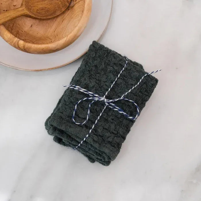 Stack of dark green towels tied with twine on a light surface with a wooden spoon and bowl in the background.