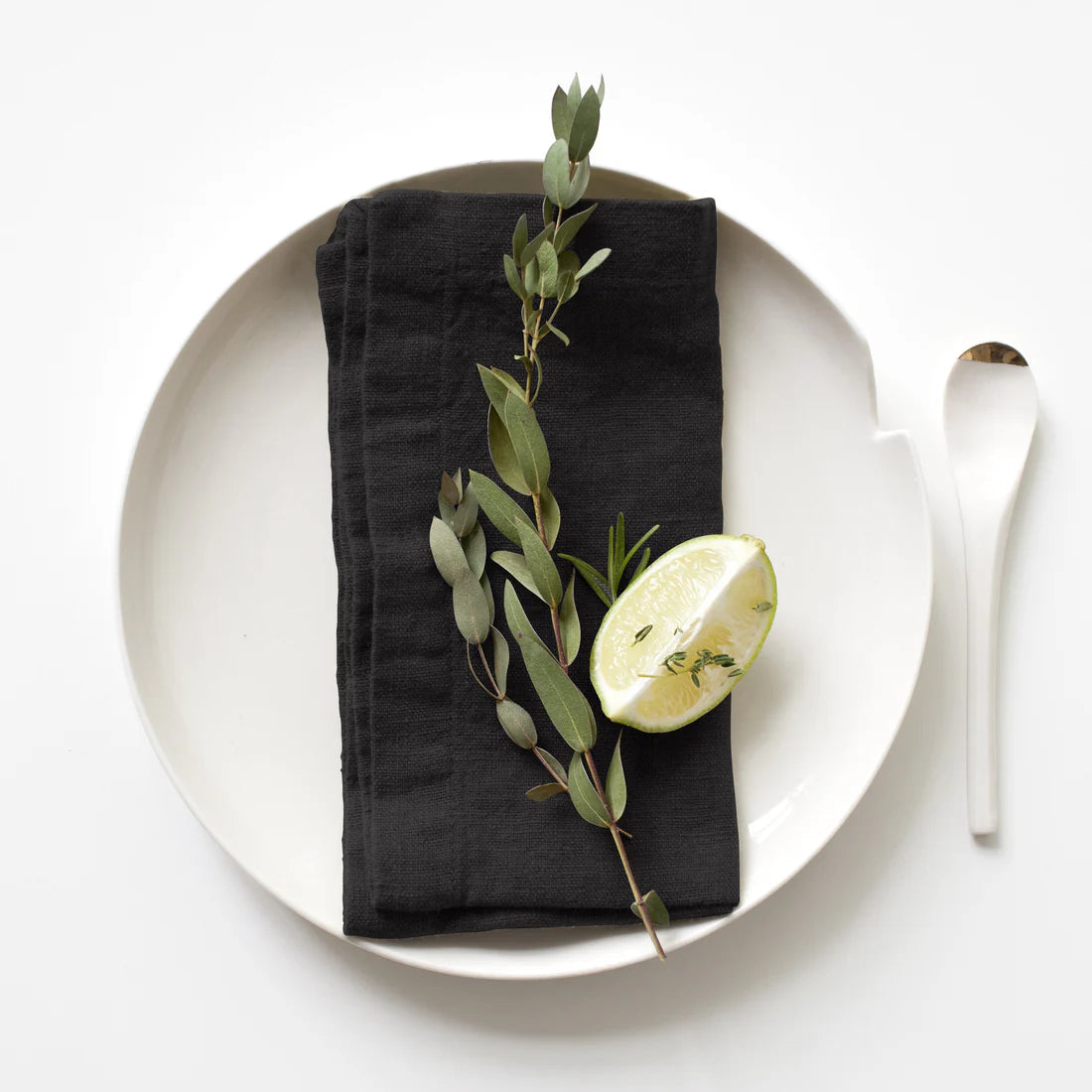 A black linen napkin is displayed on a white plate with a white spoon and adorned with a green plant and a slice of lemon, representing a sophisticated table setting.
