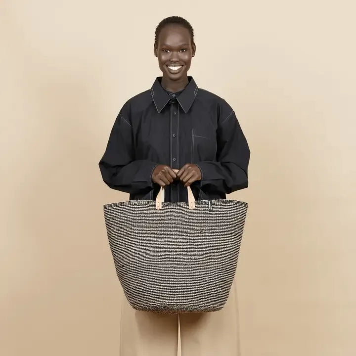 A smiling woman stands against a plain beige studio background, wearing a black collared shirt and holding a large woven tote bag with a gray-and-white textured body and light tan handles.
