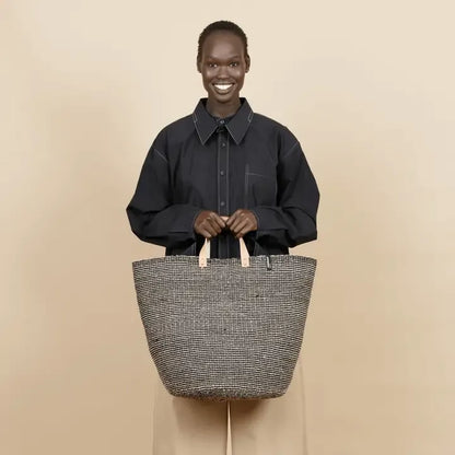 A smiling woman stands against a plain beige studio background, wearing a black collared shirt and holding a large woven tote bag with a gray-and-white textured body and light tan handles.