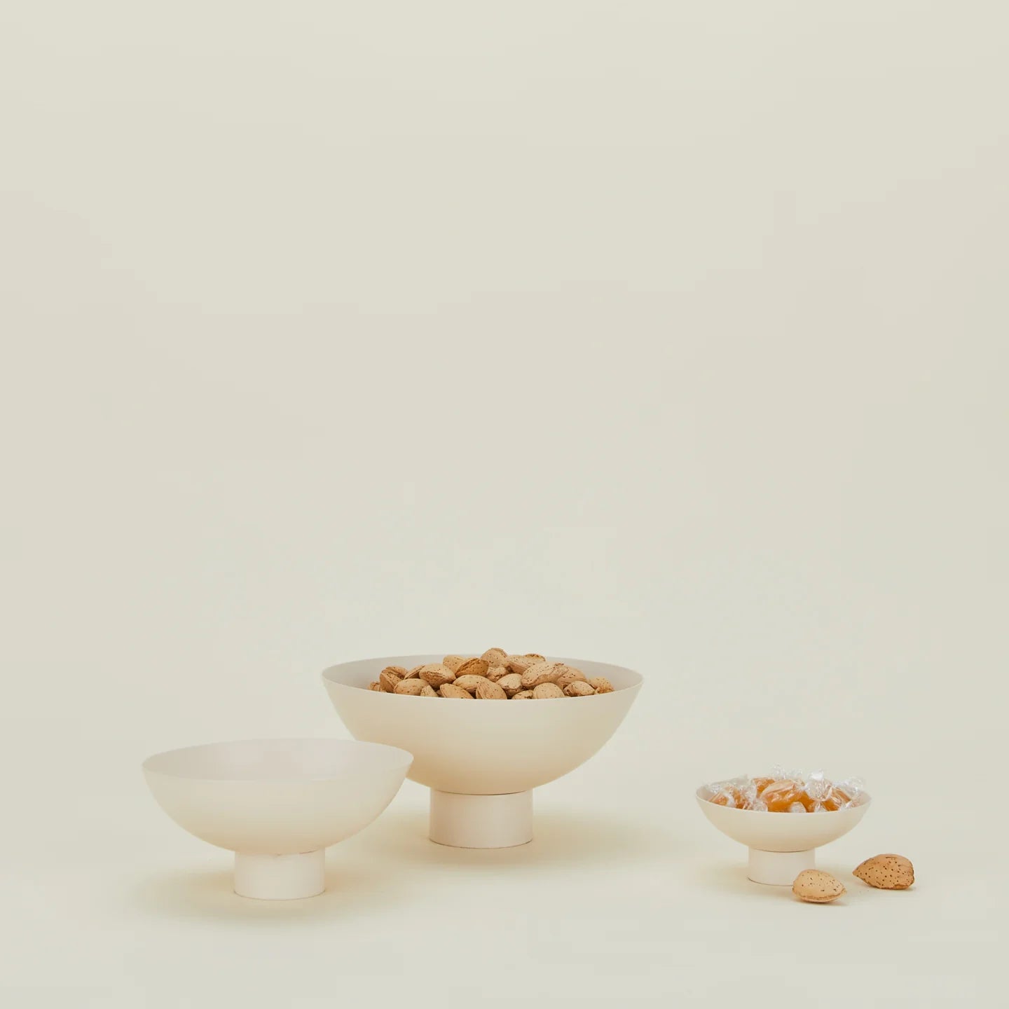 A set of three beige, powder-coated stainless steel footed bowls of various sizes, with one containing nuts displayed in the foreground.