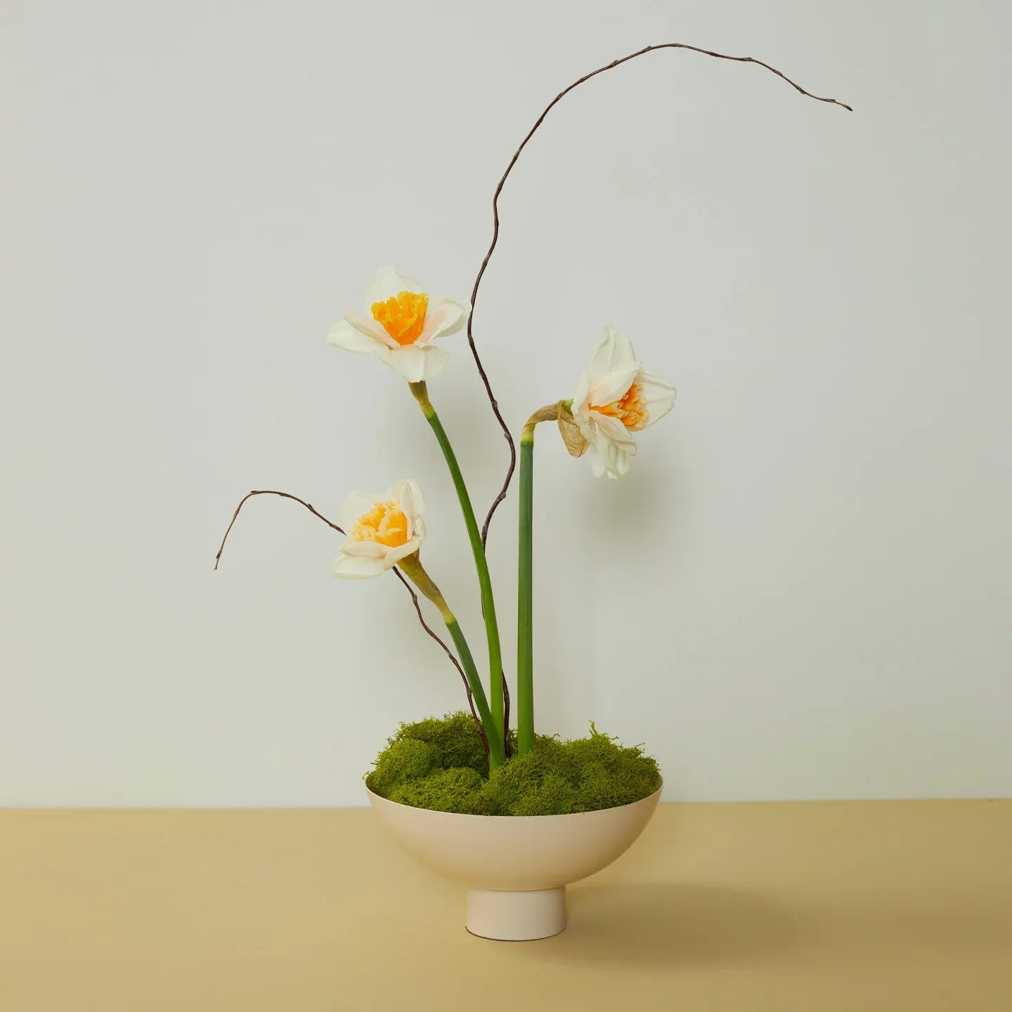 Three white flowers with yellow centers in a white bowl on a beige surface with a light gray background