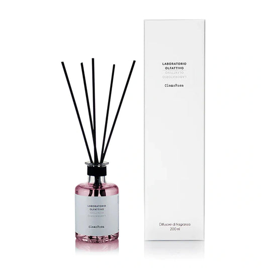 A reed diffuser bottle with pink liquid and black reeds stands next to its tall white packaging box on a clean white background