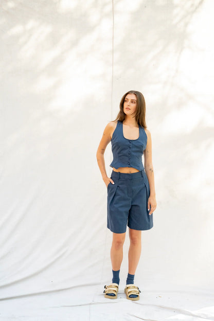 A woman modeling a matching slate blue two-piece set consisting of a sleeveless crop top and shorts, standing against a white draped backdrop with soft leaf shadows.
