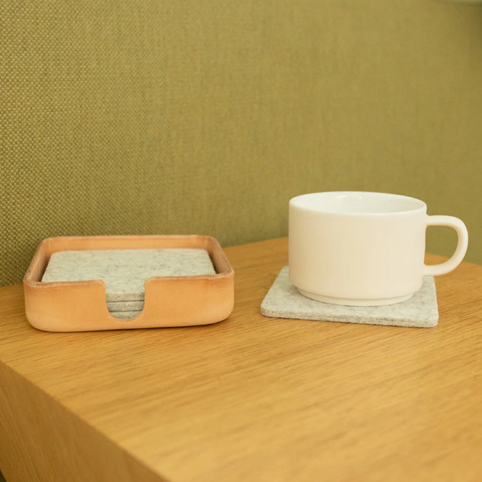 White mug on a coaster with a leather tray containing coasters on a wooden surface.
