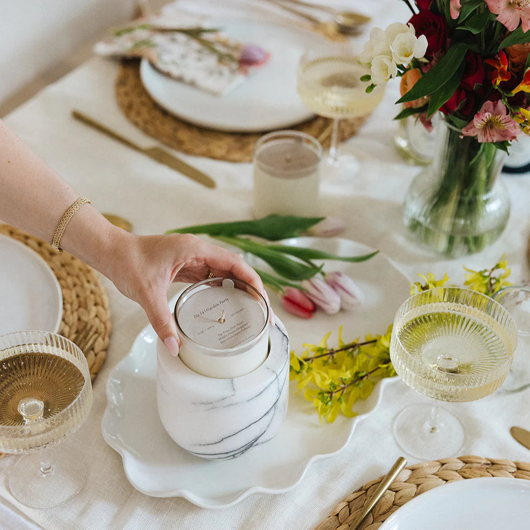 Person holding a marble-patterned candle on a elegantly set table with flowers and glasses.