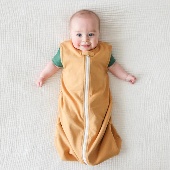 Baby wearing a yellow sleep sacwith a white zipper on a white blanket.