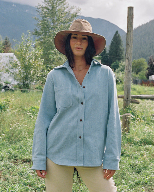Woman wearing a light blue shirt and wide-brimmed hat in a natural setting with mountains in the background.
