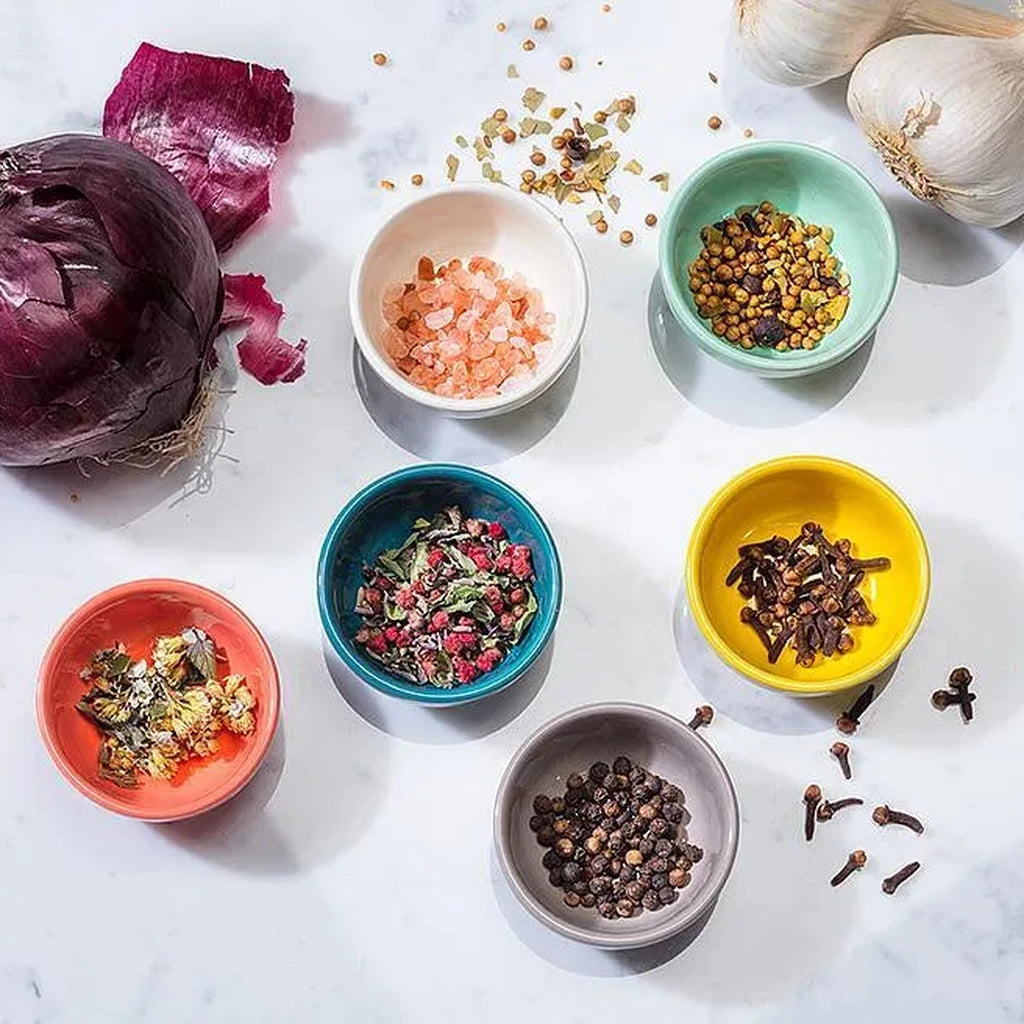 Colorful bowls of spices and herbs on a white surface with a red onion and garlic.