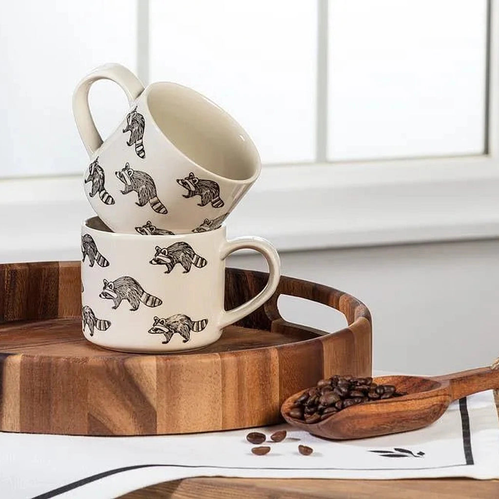 Two stacked ceramic mugs with a black raccoon pattern on a cream background, displayed on a wooden tray with coffee beans and a wooden spoon in the foreground.