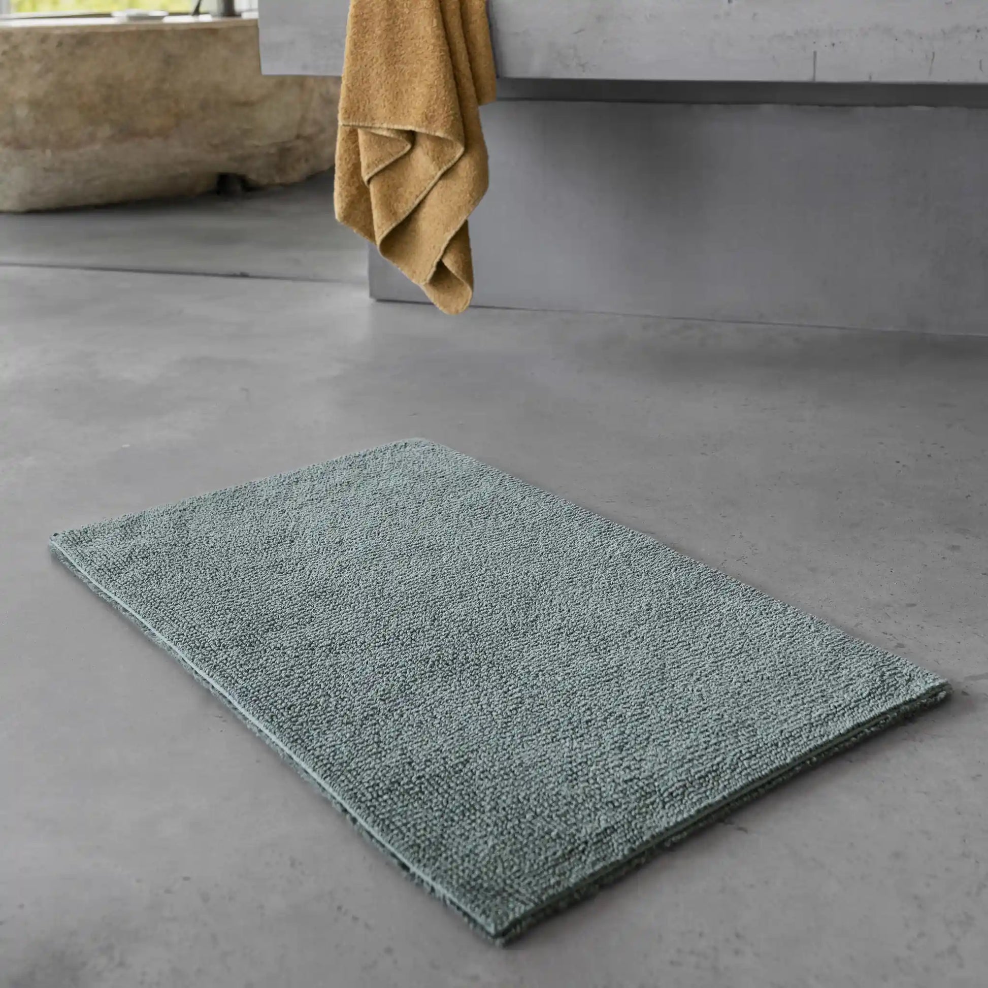 Green bath mat on a gray bathroom floor with a towel hanging in the background.