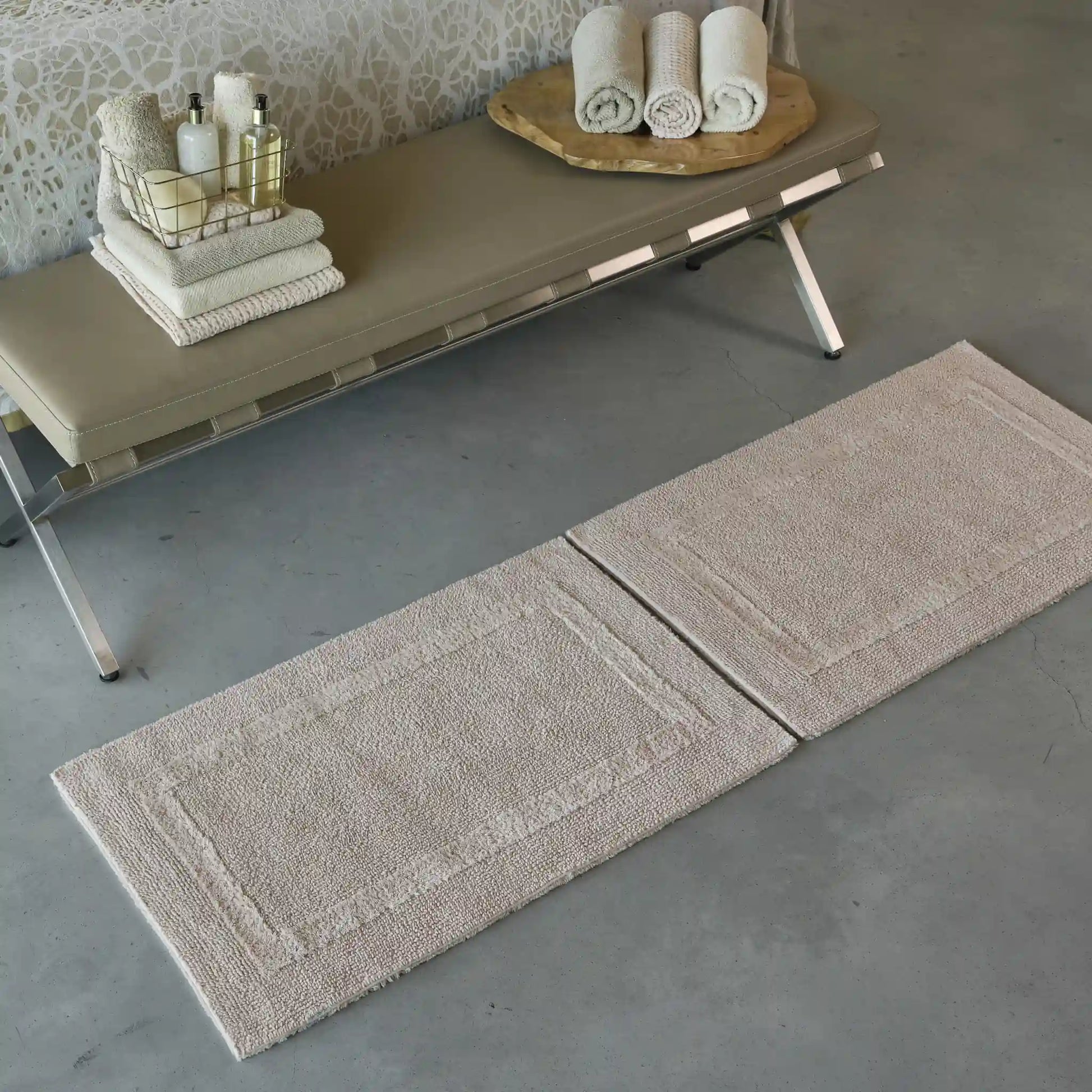Two beige bath mats on a gray floor with a bench and towels in the background.