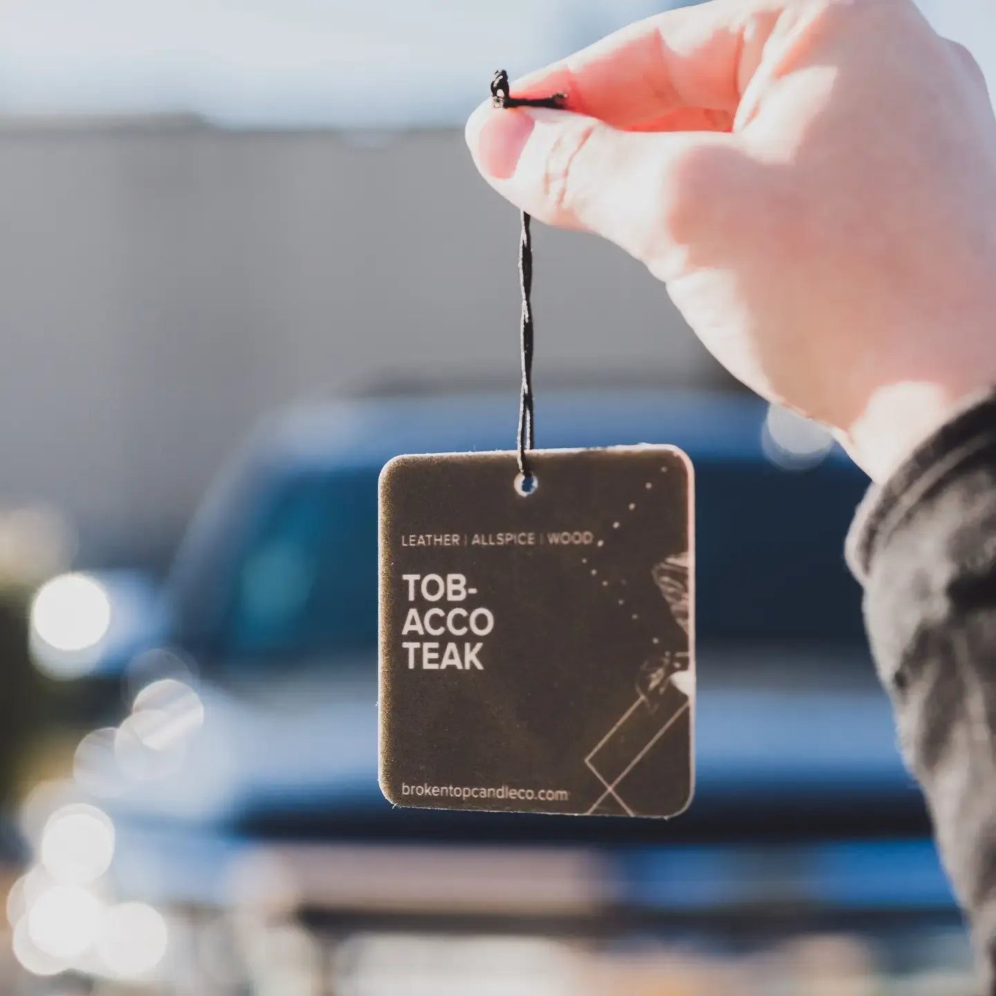 A hand holds a square dark brown car air freshener tag labeled 'TOBACCO TEAK' against a blurred outdoor background with a vehicle.