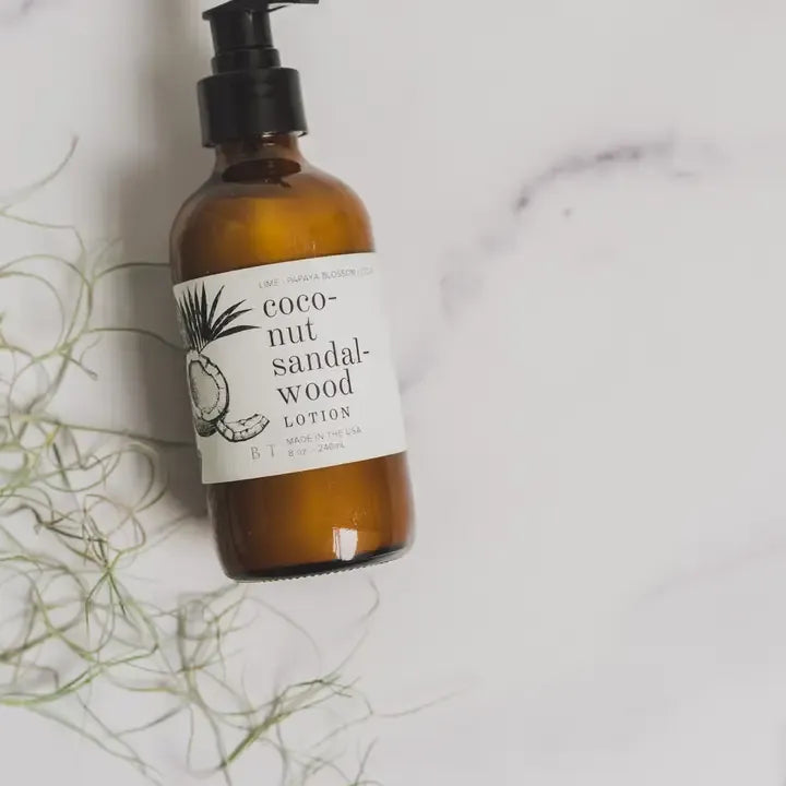 An amber glass pump bottle of coconut sandalwood lotion with a black pump and a white label, styled on a white marble surface with dried green plant stems.