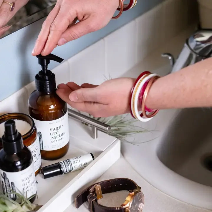A person's hands pump a bottle of Saguaro Cactus Lotion in a bathroom vanity tray with other skincare and lifestyle items.