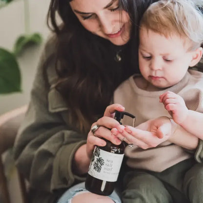 A woman and a young child are sitting together indoors, with the woman holding a dark bottle labeled 'sea salt lotion' and pumping soap into the child's hand.