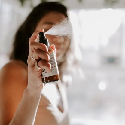 A woman holds a brown glass spray bottle with a black pump, spraying a fine mist toward the camera. The bottle has a white label with black text, and the background is a bright, softly blurred indoor setting with warm light.