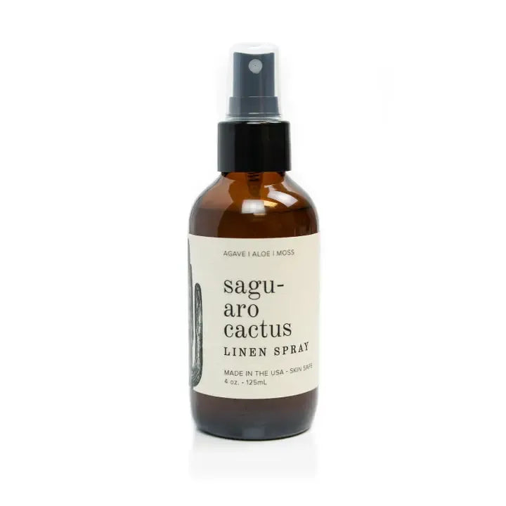 An amber glass spray bottle of saguaro cactus linen spray with a black pump and a cream label, set against a plain white background.