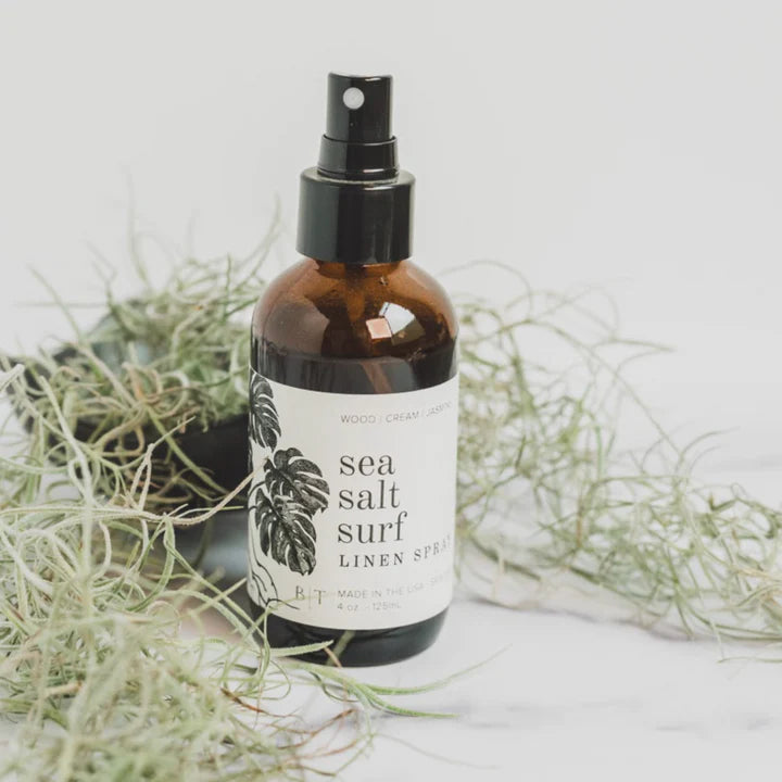 A linen spray bottle with a label reading 'sea salt surf LINEN SPRAY', surrounded by what appears to be dried grasses or plant material.
