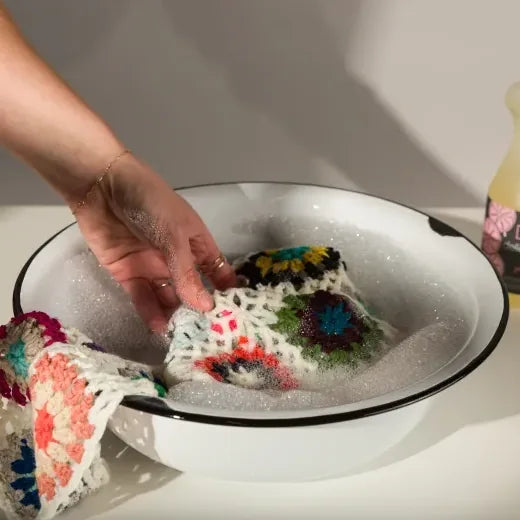 Colorful crocheted item being washed in a white bowl with soapy water.