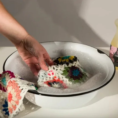Colorful crocheted item being washed in a white bowl with soapy water.