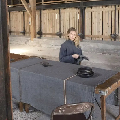 A woman in a dark blue-gray tunic stands behind a rustic wooden table draped with a matching gray linen cloth, holding a small dark object in a barn-like setting with wooden walls.