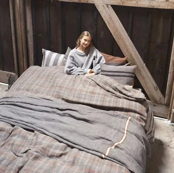 Woman sitting on a bed with plaid and gray bedding in a rustic setting.