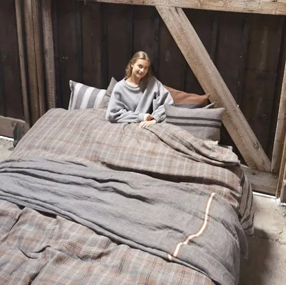 Woman sitting on a bed with plaid and gray bedding in a rustic setting.
