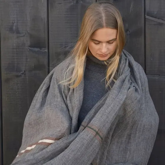 A woman with blonde hair wrapped in a large gray linen blanket over a dark gray turtleneck sweater, standing against a dark wooden plank background.