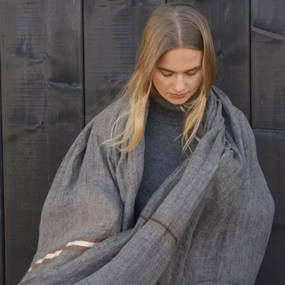 A woman with blonde hair wrapped in a large gray linen blanket over a dark gray turtleneck sweater, standing against a dark wooden plank background.