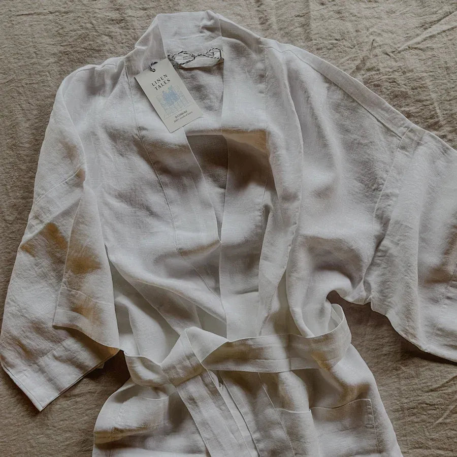 A white linen bathrobe with a tie belt, laid flat on a textured beige fabric background. 