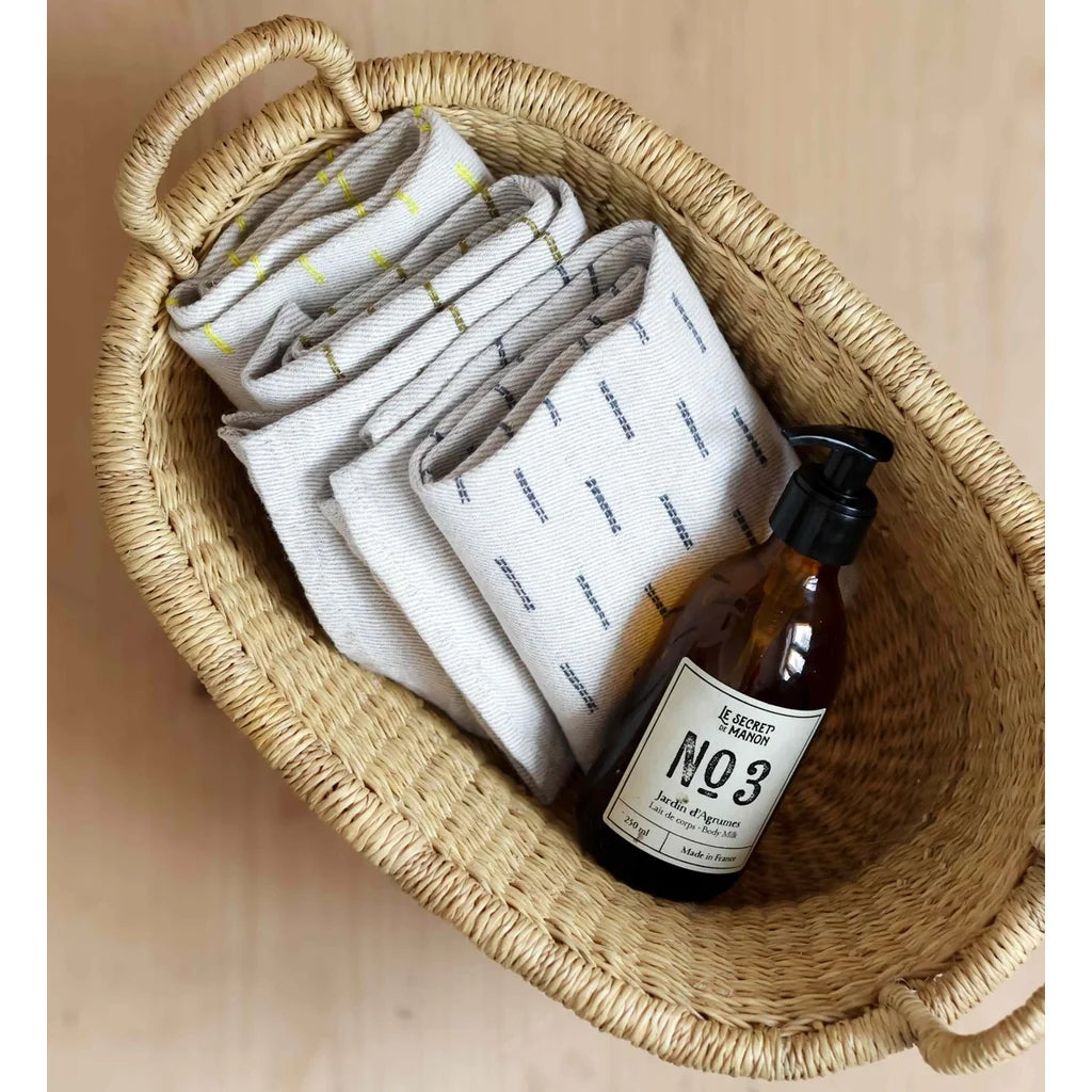 A woven oval basket containing folded white towels with blue and yellow stripes, alongside an amber glass pump bottle.