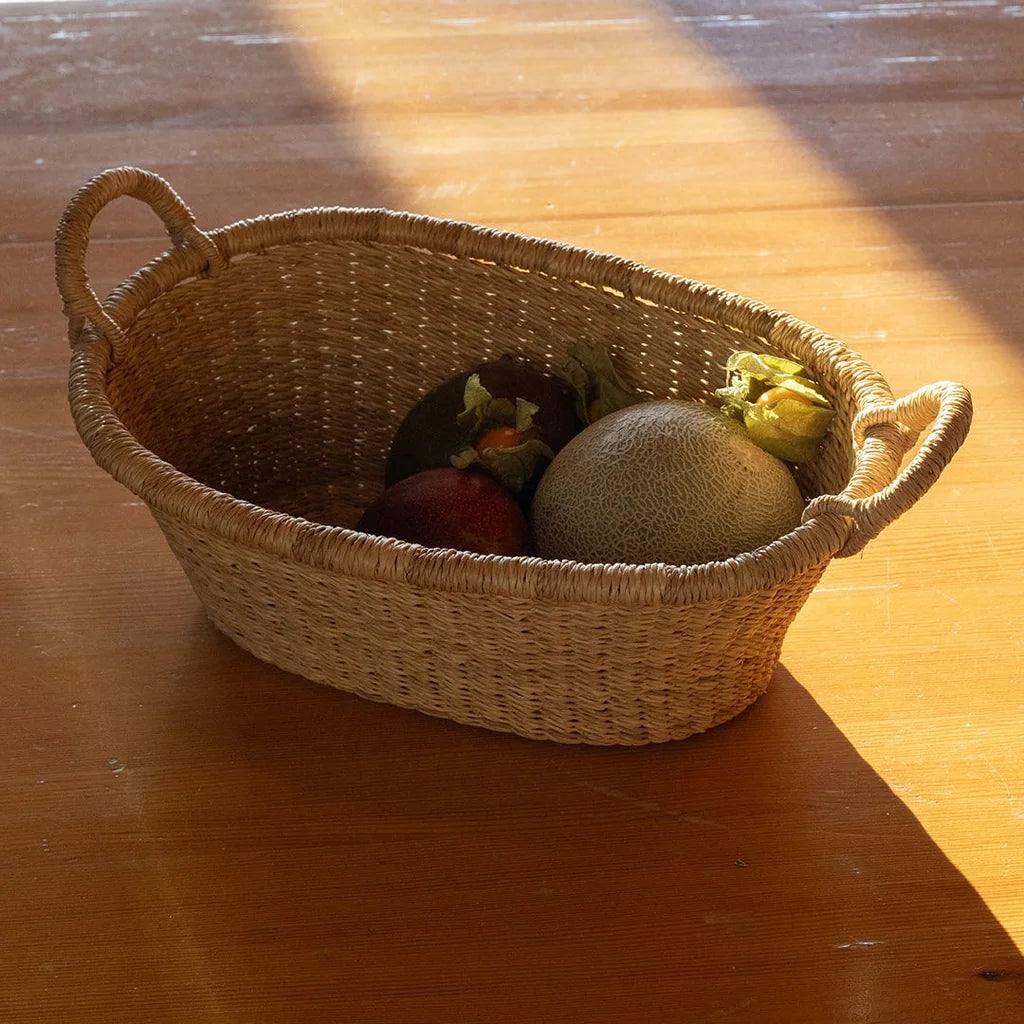 A natural woven straw basket with two side handles sits on a wooden table, holding fruit and vegetables under warm sunlight.
