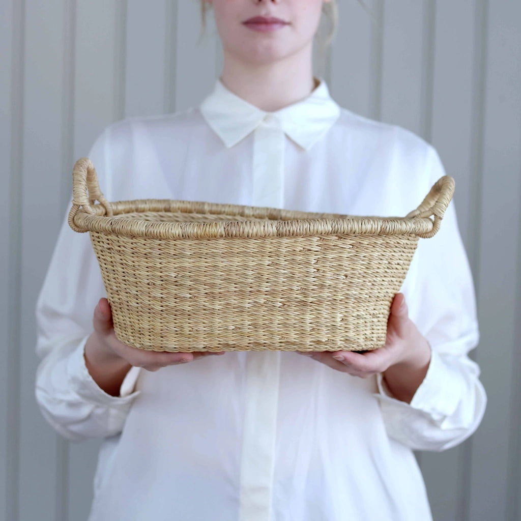 A person in a white collared shirt holds a natural woven straw basket with side handles against a light gray paneled background.