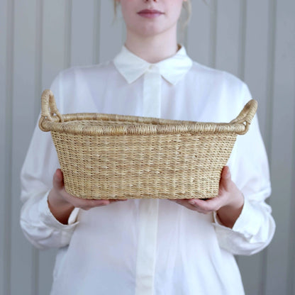 A person in a white collared shirt holds a natural woven straw basket with side handles against a light gray paneled background.