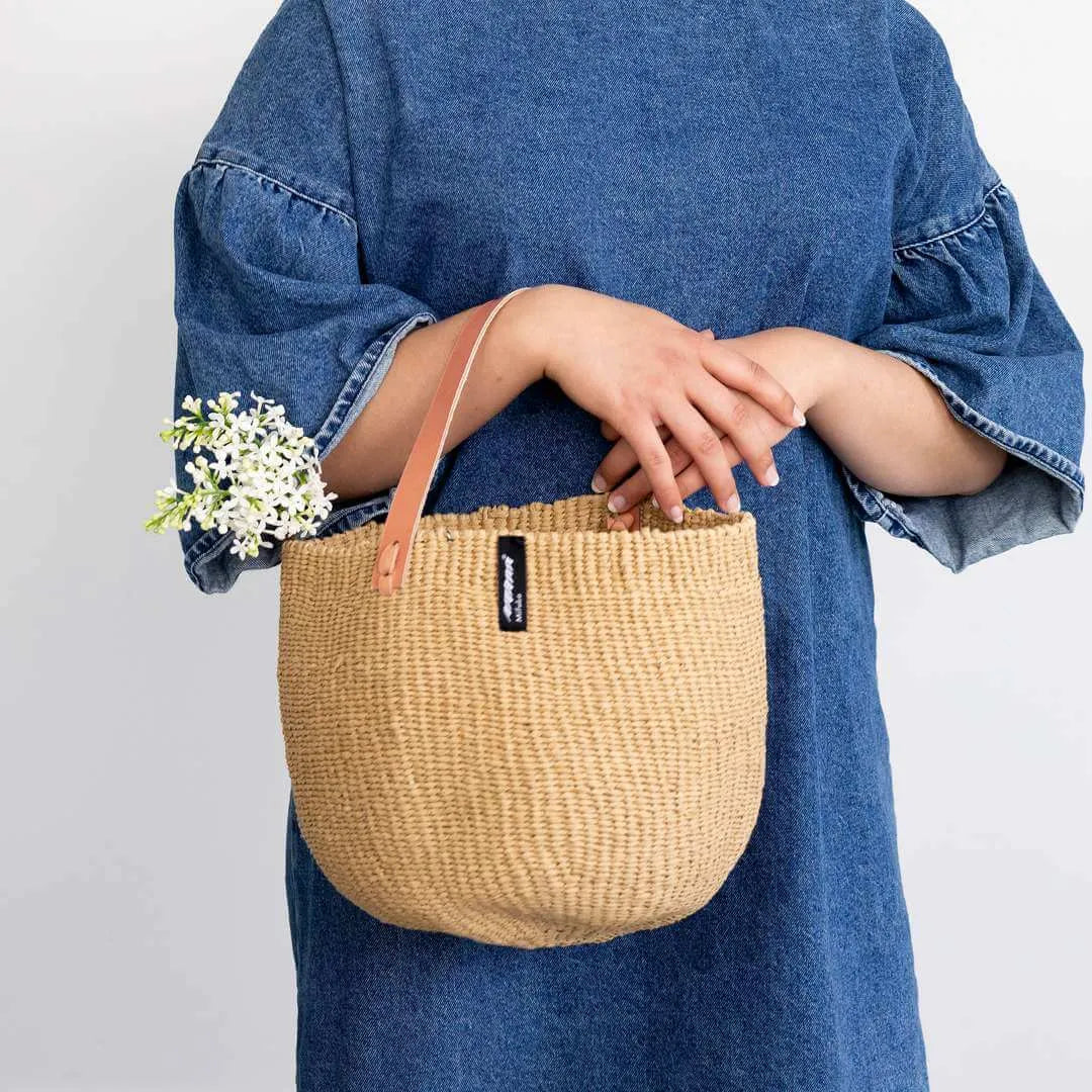 Person holding a woven bag with a small plant, wearing a blue denim outfit on a plain background