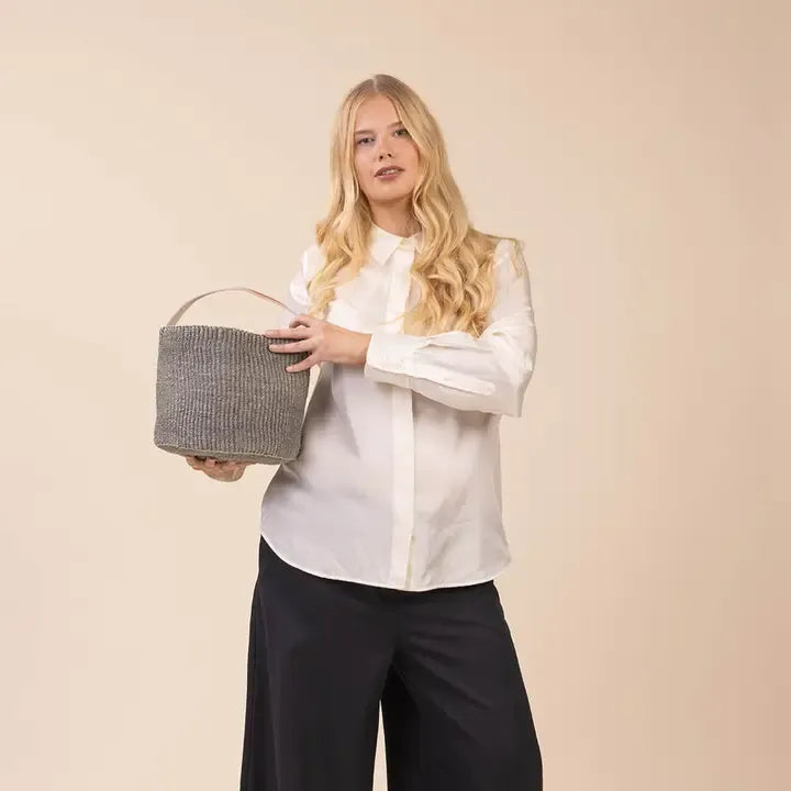 A blonde woman wearing a white long-sleeve blouse and black wide-leg pants holds a gray woven basket bag with a light brown handle against a solid beige studio background.