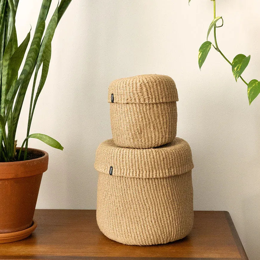 Two stacked round woven storage baskets in a natural beige jute material, displayed on a wooden table next to a potted snake plant against a plain off-white wall.