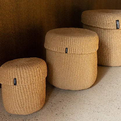 Three round woven storage baskets with lids in natural straw color, arranged on a speckled floor against a dark wood background.