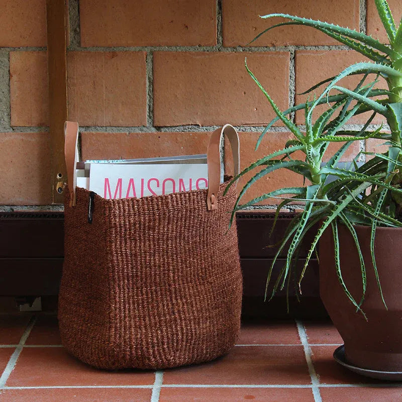 Brown woven basket with a plant and a magazine against a brick wall.