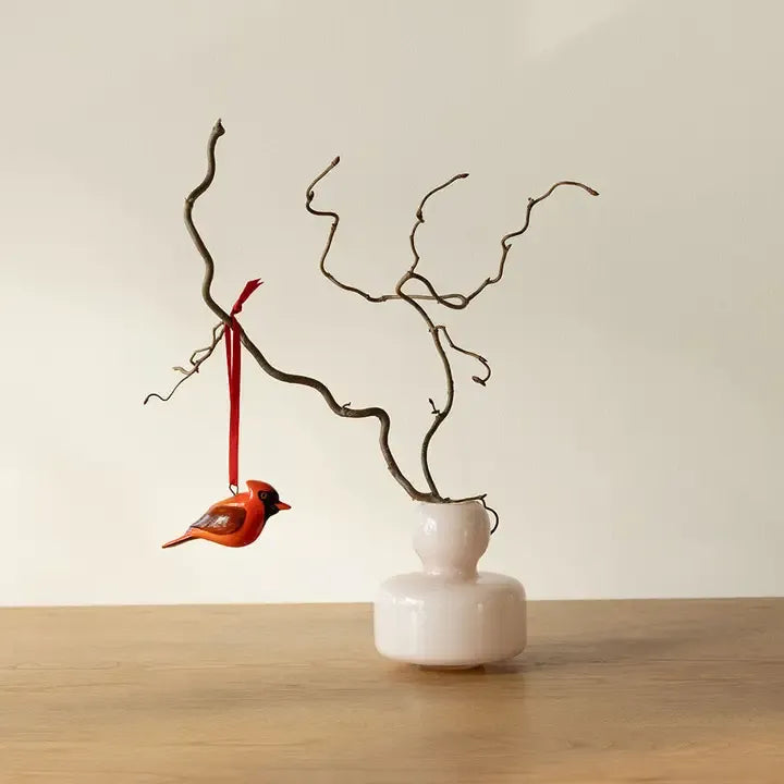 Decorative branch with a small bird ornament hanging from it, placed in a white vase on a wooden surface.
