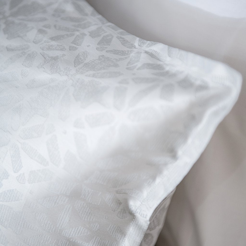 A close-up of a white pillowcase with a subtle tonal floral pattern, resting on a matching white pillow.