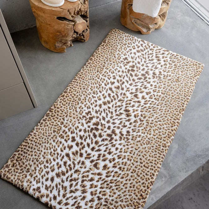 Abyss & Habidecor Feline leopard print bath rug on a grey concrete floor beside wooden stools holding white slippers and a towel.