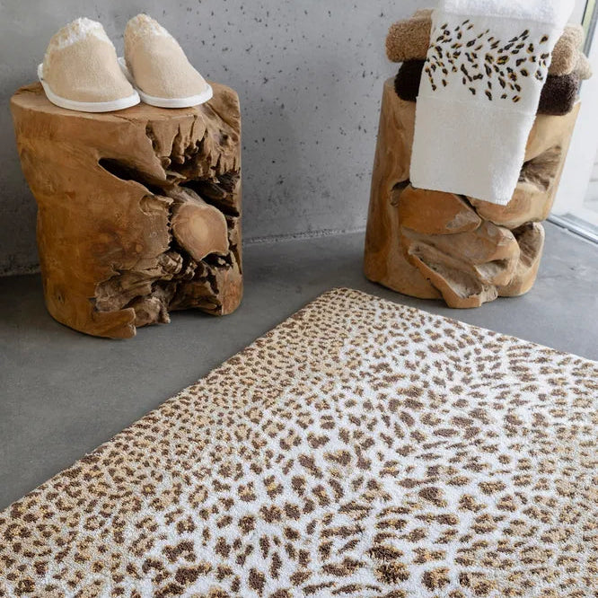 Closer view of the Feline leopard print bath rug on a concrete floor next to wooden stools with slippers and a towel.