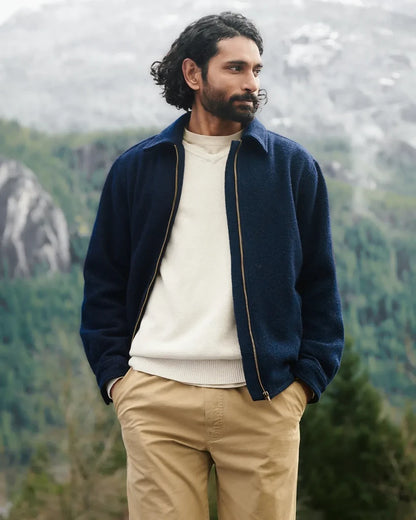 A man wearing a navy blue zip-up fleece jacket over a cream knit sweater and khaki pants, standing outdoors with a blurred mountain and forest background.