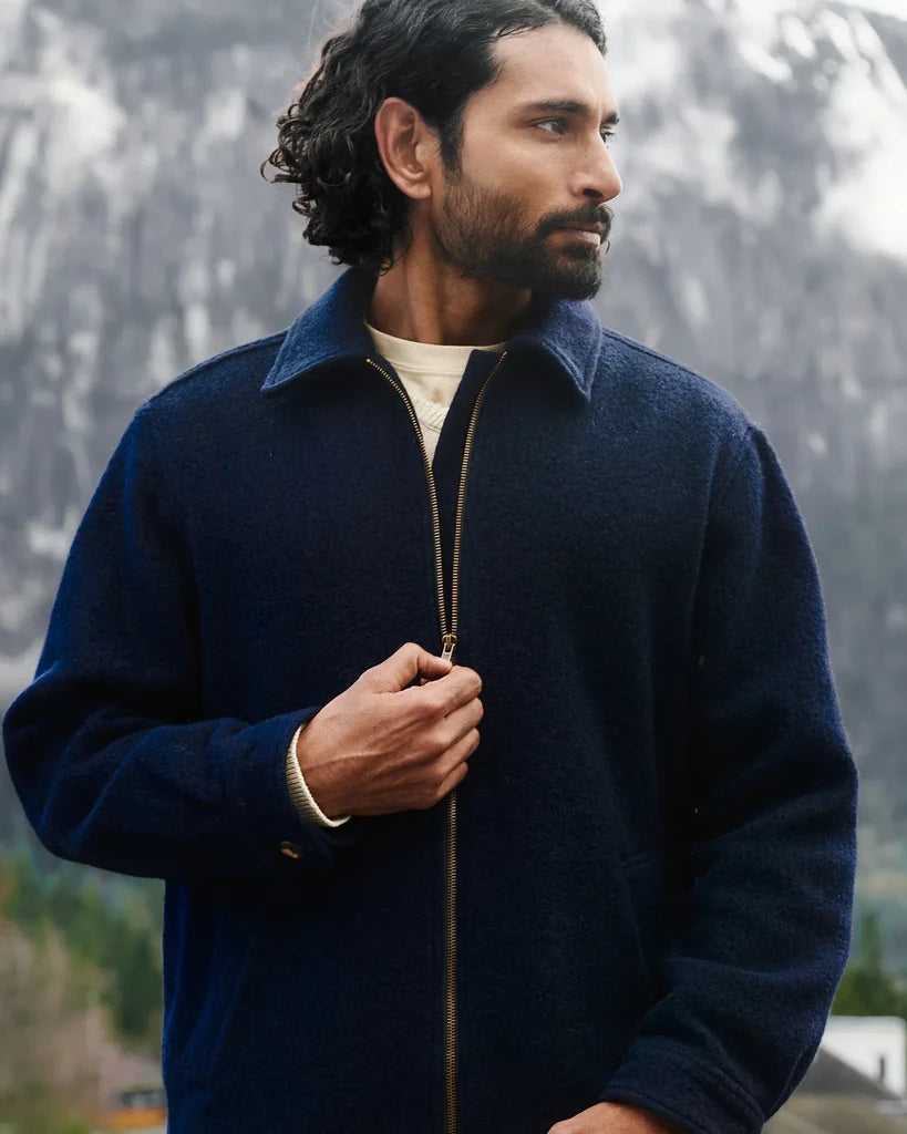 A man wearing a dark navy blue zip-up fleece jacket over a cream t-shirt, standing outdoors with a blurred mountain background.