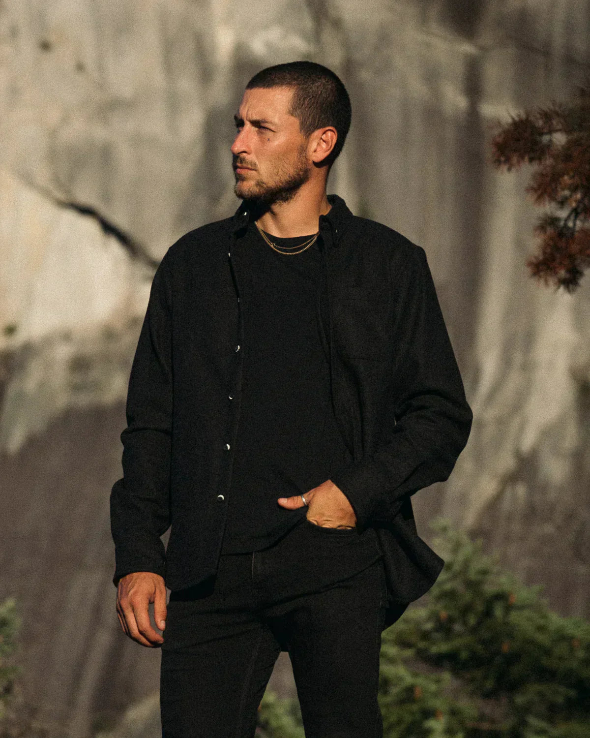 A man wearing a black button-up shirt and black pants stands outdoors against a rocky, sunlit cliff face.