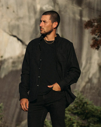 A man wearing a black button-up shirt and black pants stands outdoors against a rocky, sunlit cliff face.